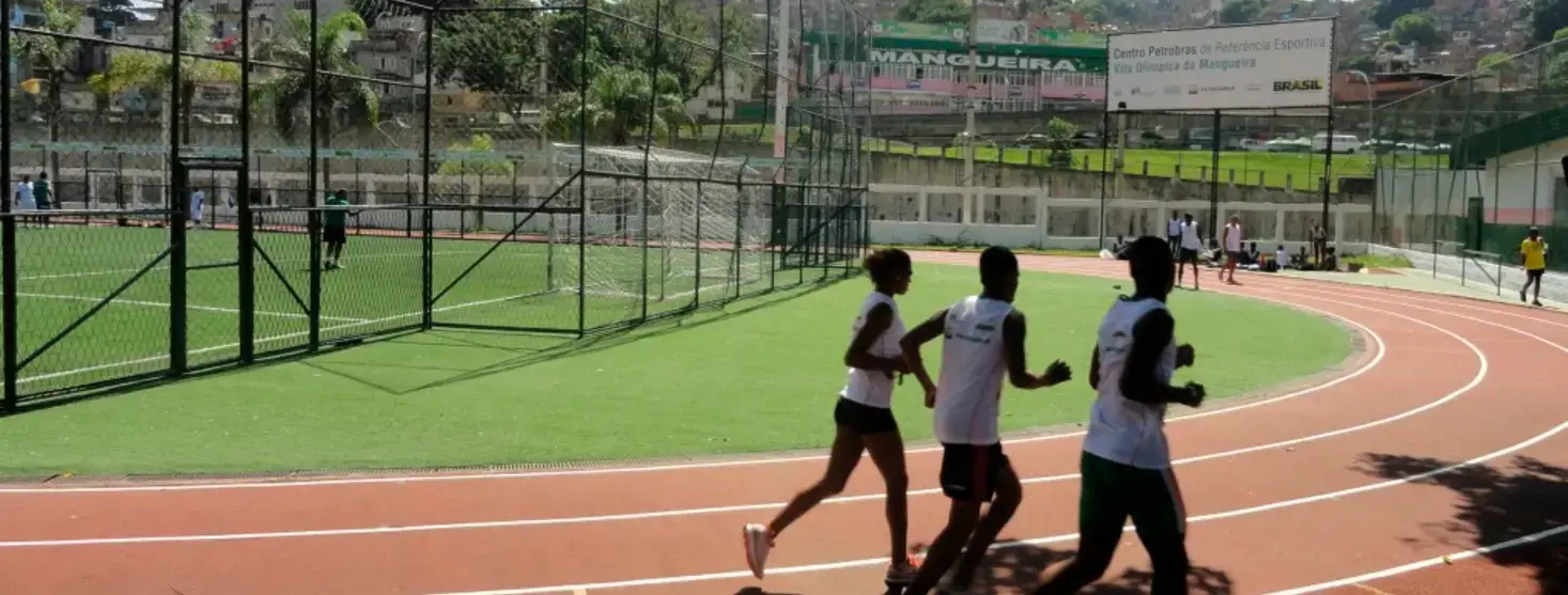 Três jovens correm numa pista de atletismo vermelha ao lado de um campo de grama sintética cercado, com a placa da Vila Olímpica da Mangueira ao fundo