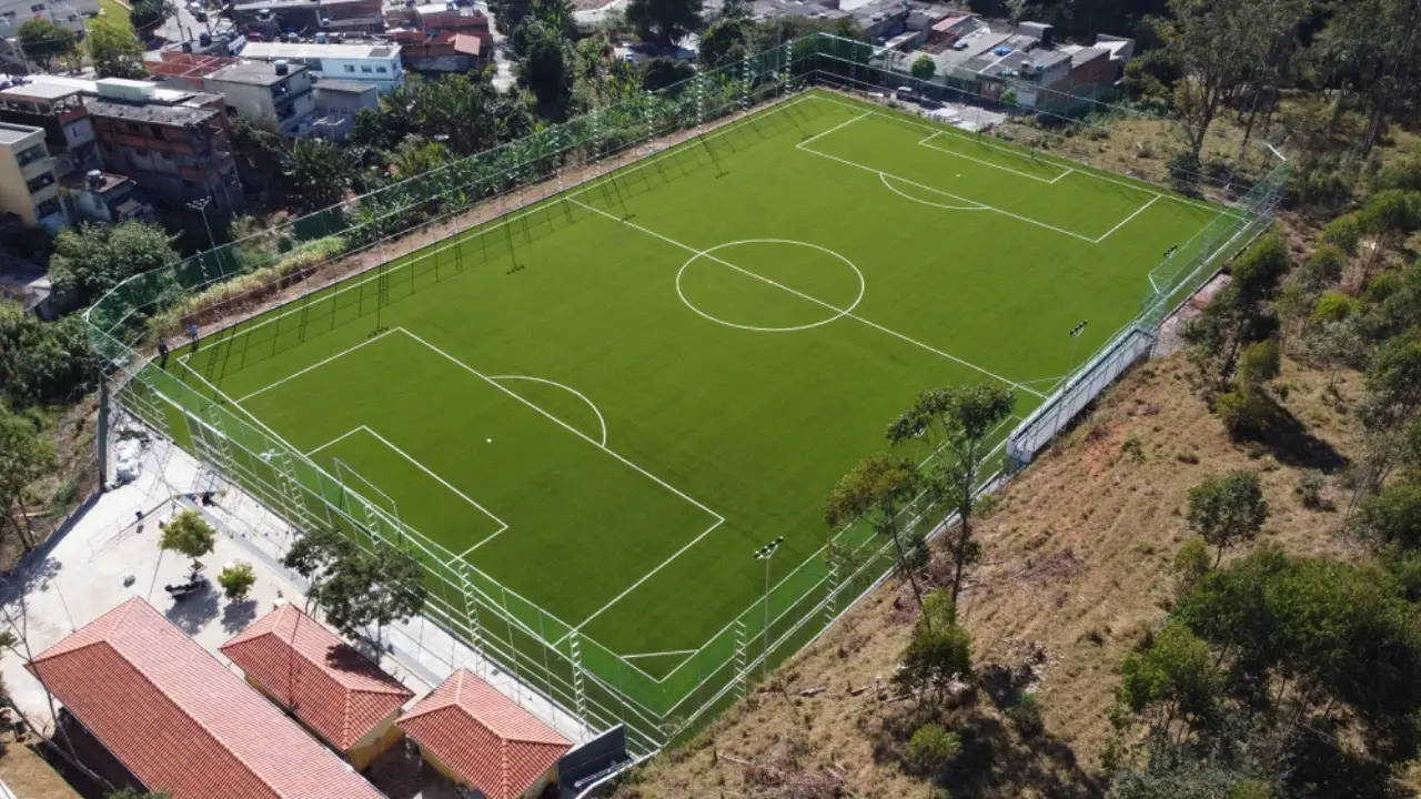 Vista aérea de um campo de futebol de grama sintética cercado por alambrado, entre casas e um morro com vegetação