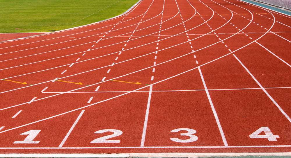 Saída de uma pista de atletismo sintética vermelha com as raias 1 a 4 numeradas e o gramado ao fundo