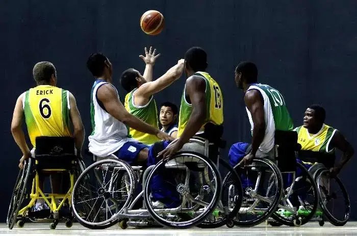 Jogadores de basquete em cadeira de rodas, de uniformes amarelo e branco, disputam o rebote em ginásio coberto