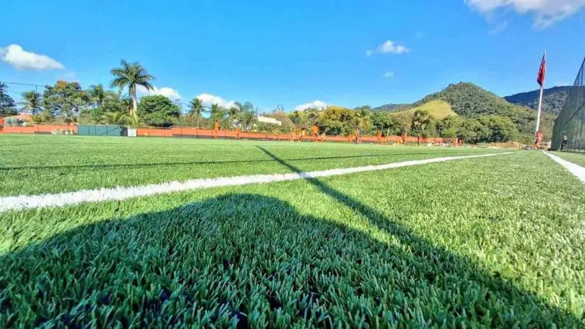 Campo de grama sintética visto rente ao chão, com linha branca de marcação, jogadores de laranja ao fundo e morros sob céu azul