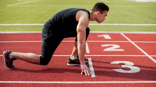 Atleta agachado em posição de largada sobre a linha branca de uma pista sintética vermelha, com os números das raias pintados no piso