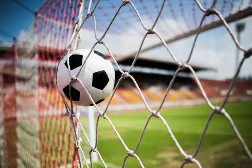 Bola de futebol estufando a rede do gol, com o gramado e a arquibancada de um estádio ao fundo