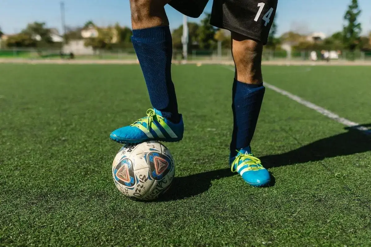 Jogador com chuteira azul pisa sobre a bola num campo de grama sintética com linha branca de marcação