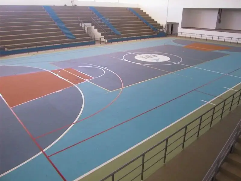 Ginásio coberto com piso sintético azul, cinza e laranja, linhas de futsal, basquete e vôlei e arquibancada ao fundo