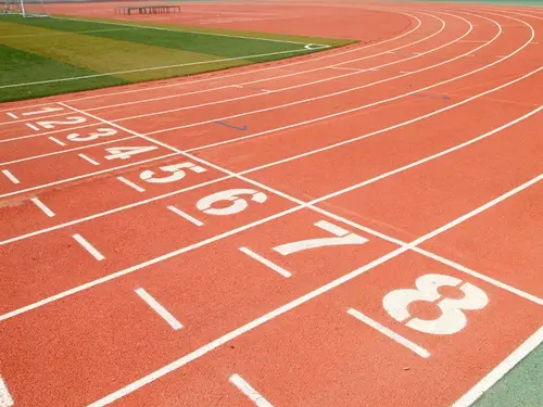 Saída de curva de uma pista sintética vermelha com as raias numeradas de 1 a 8 e o gramado ao fundo