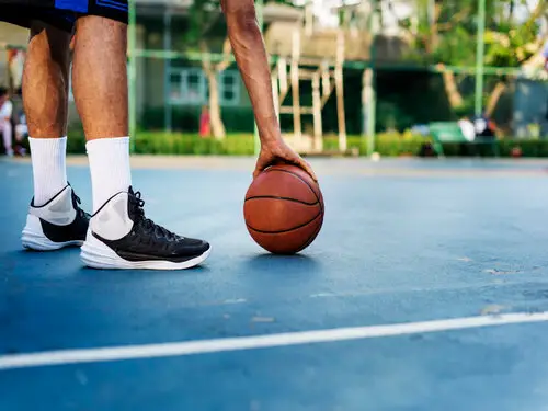 Jogador de tênis preto e meias brancas segura a bola de basquete sobre uma quadra aberta de piso azul com linha branca, com árvores ao fundo