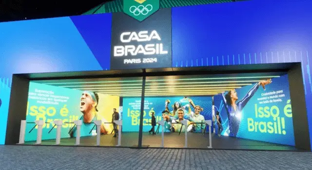 Projeção em 3D da fachada da Casa Brasil Paris 2024, com letreiro luminoso, aros olímpicos e painéis de atletas com a frase Isso é Brasil