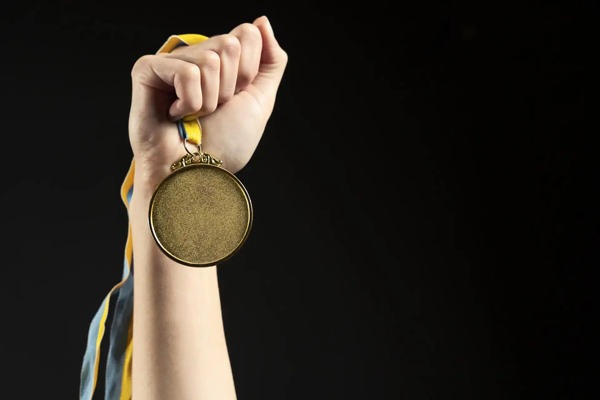 Mão erguida segurando uma medalha dourada com fita amarela e azul, sobre fundo preto
