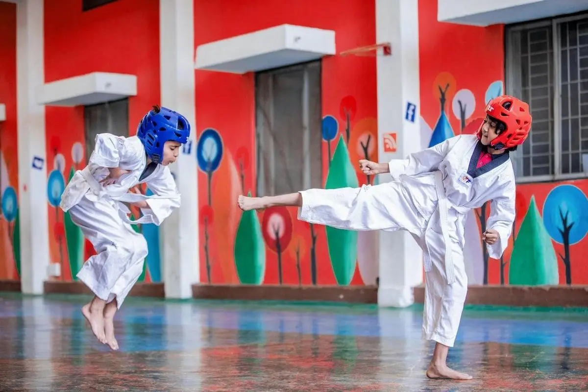 Duas crianças de dobok branco e capacete treinam taekwondo, uma chutando e outra saltando, diante de um muro pintado de árvores coloridas