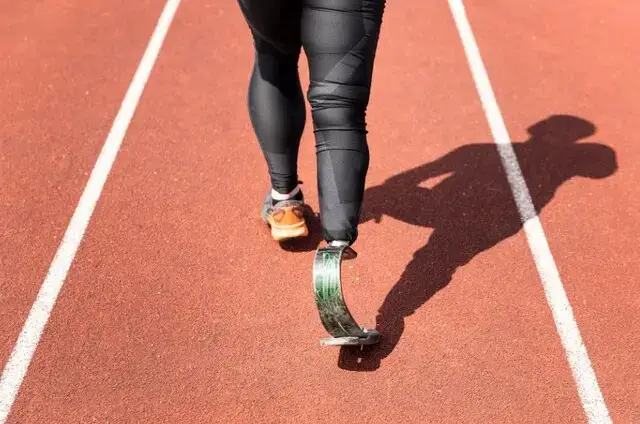 Atleta com prótese de corrida caminhando sobre pista sintética vermelha de atletismo, com as linhas brancas das raias e a sombra no chão
