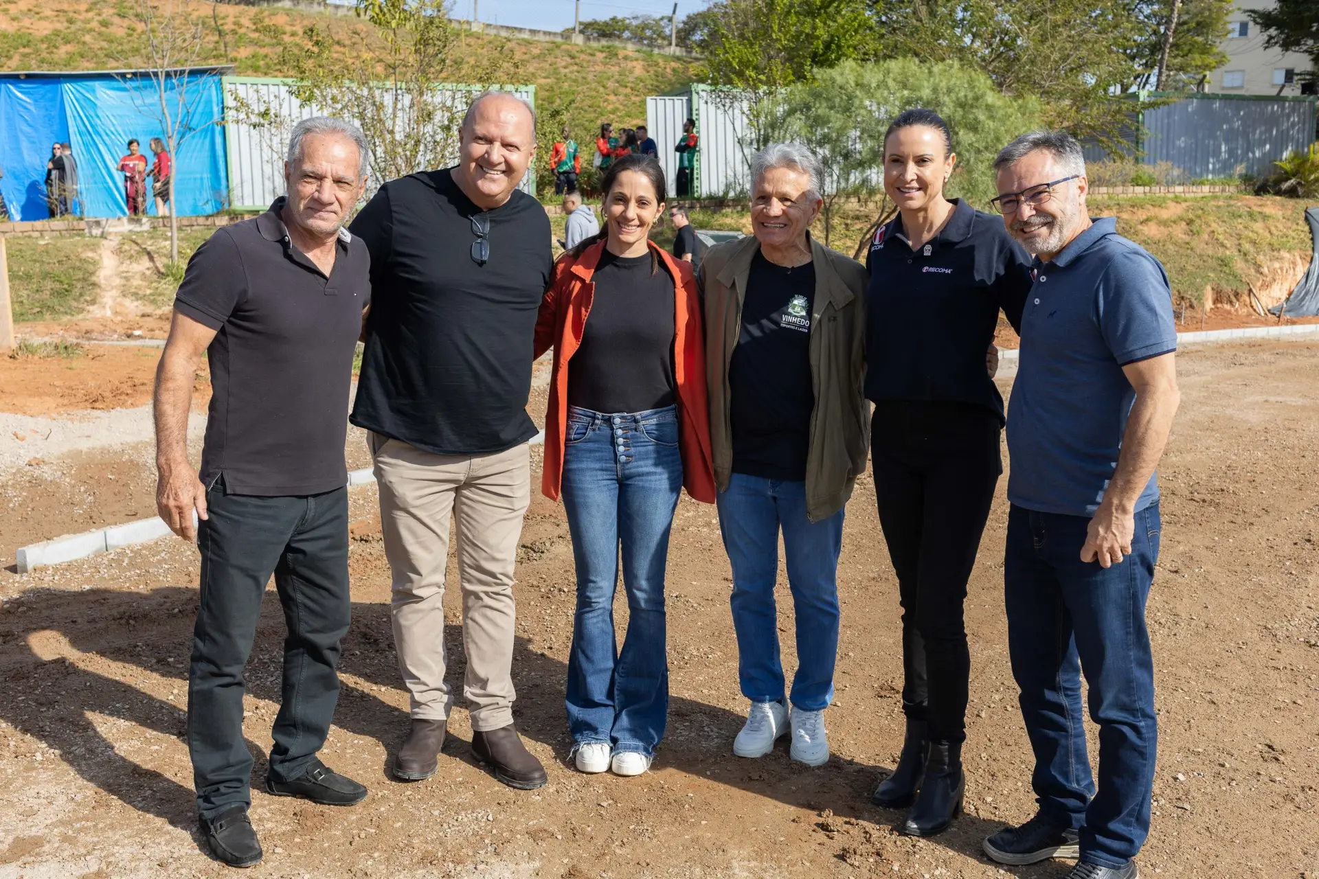 Seis pessoas lado a lado sobre terreno de terra batida em obra, uma delas com camisa da Recoma e outra com camisa de Vinhedo Esportes e Lazer, com contêineres e lona azul ao fundo