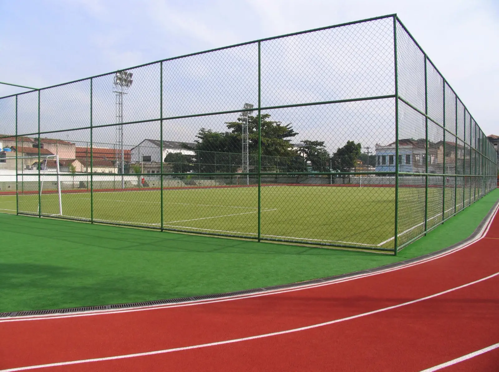 Campo de grama sintética cercado por alambrado alto de tela de aço verde, com pista de atletismo vermelha em primeiro plano e um poste de iluminação ao fundo