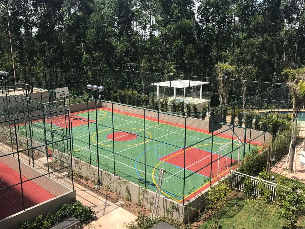 Conjunto de quadras externas com piso verde e vermelho, cercadas por alambrado de tela e postes de luz, vistas do alto entre árvores