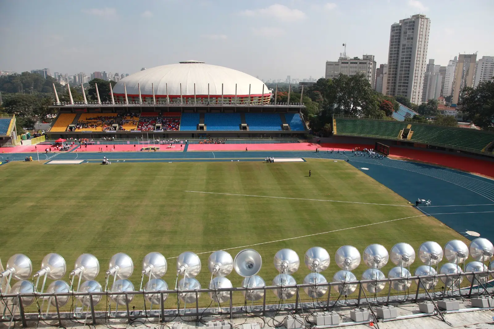 Campo de futebol num estádio com pista de atletismo azul em volta, vista de trás de uma fileira de refletores prateados, com um ginásio de cúpula branca e prédios ao fundo