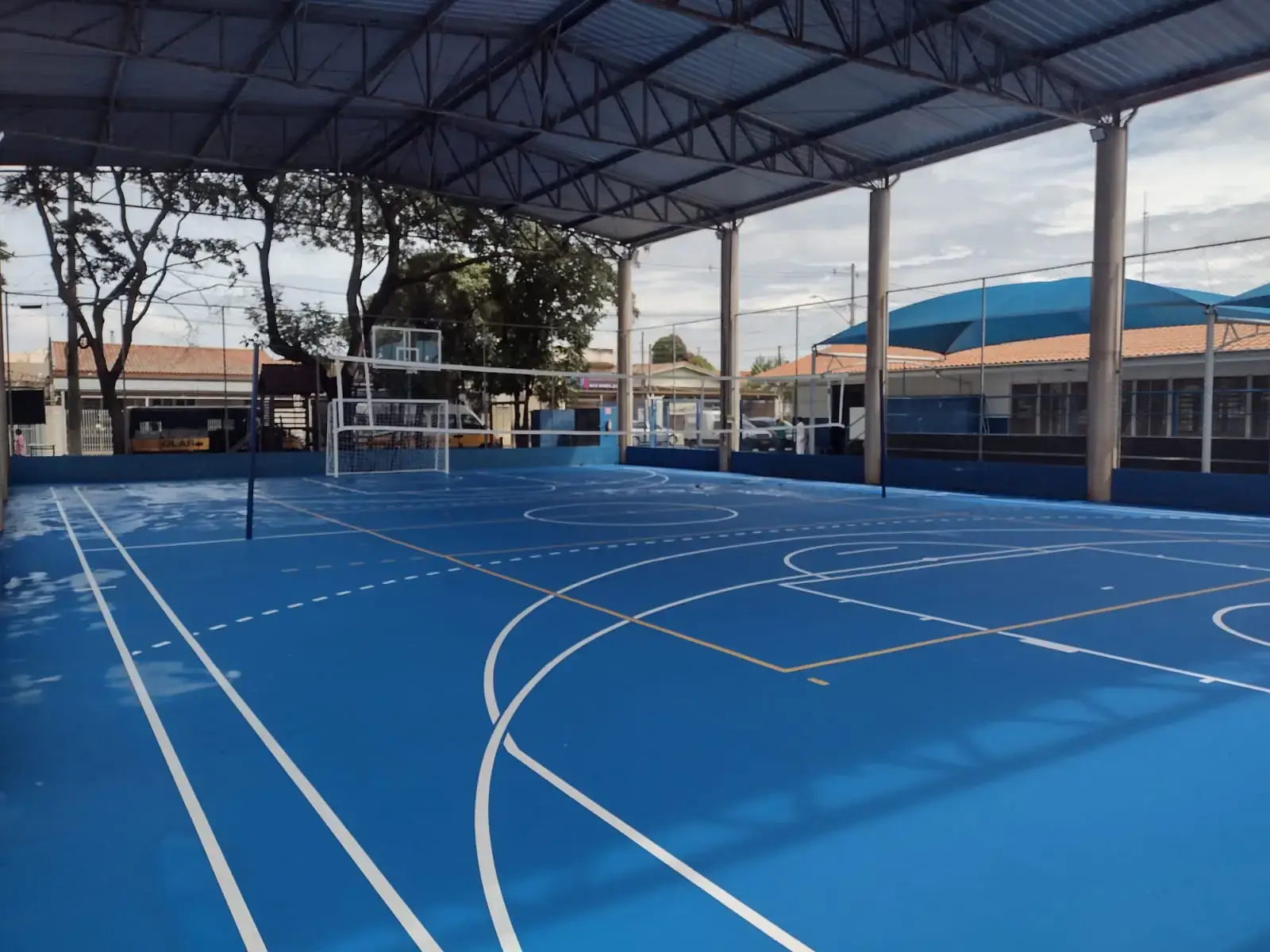 Quadra poliesportiva coberta por estrutura metálica, com piso azul liso, linhas brancas de várias modalidades e traves de futsal, aberta nas laterais para árvores e casas