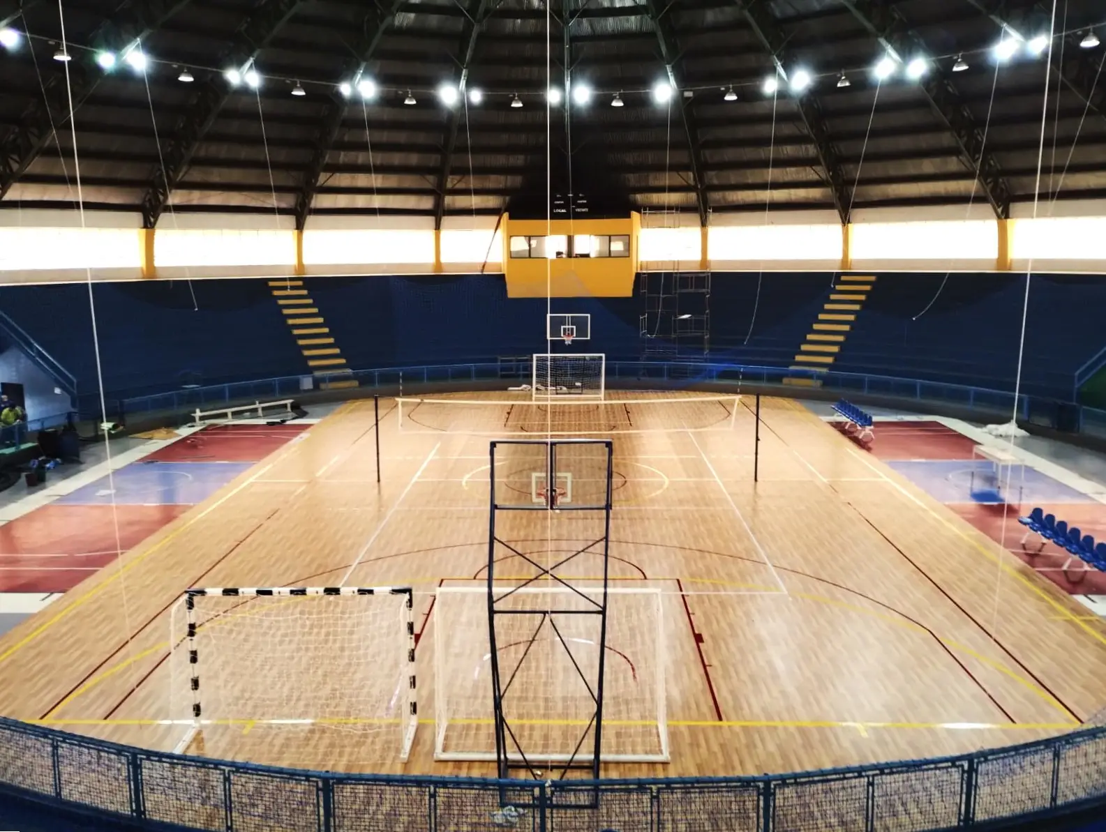 Ginásio coberto visto da arquibancada, com piso vinílico de padrão amadeirado, linhas de várias modalidades, traves de handebol e tabela de basquete
