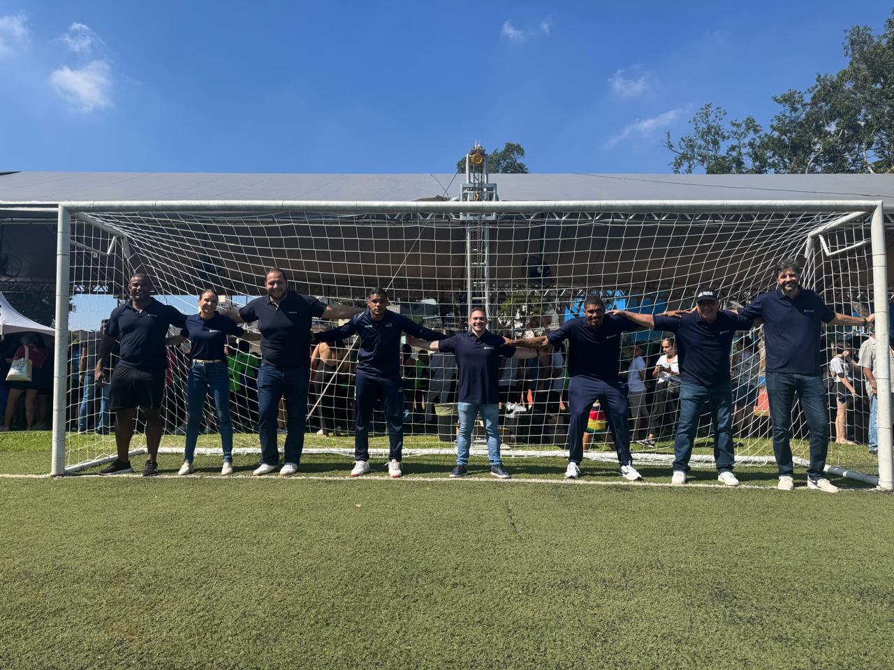 Grupo de pessoas de camisa polo azul-marinho, de braços dados diante de um gol sobre gramado sintético, com tenda de evento e público ao fundo